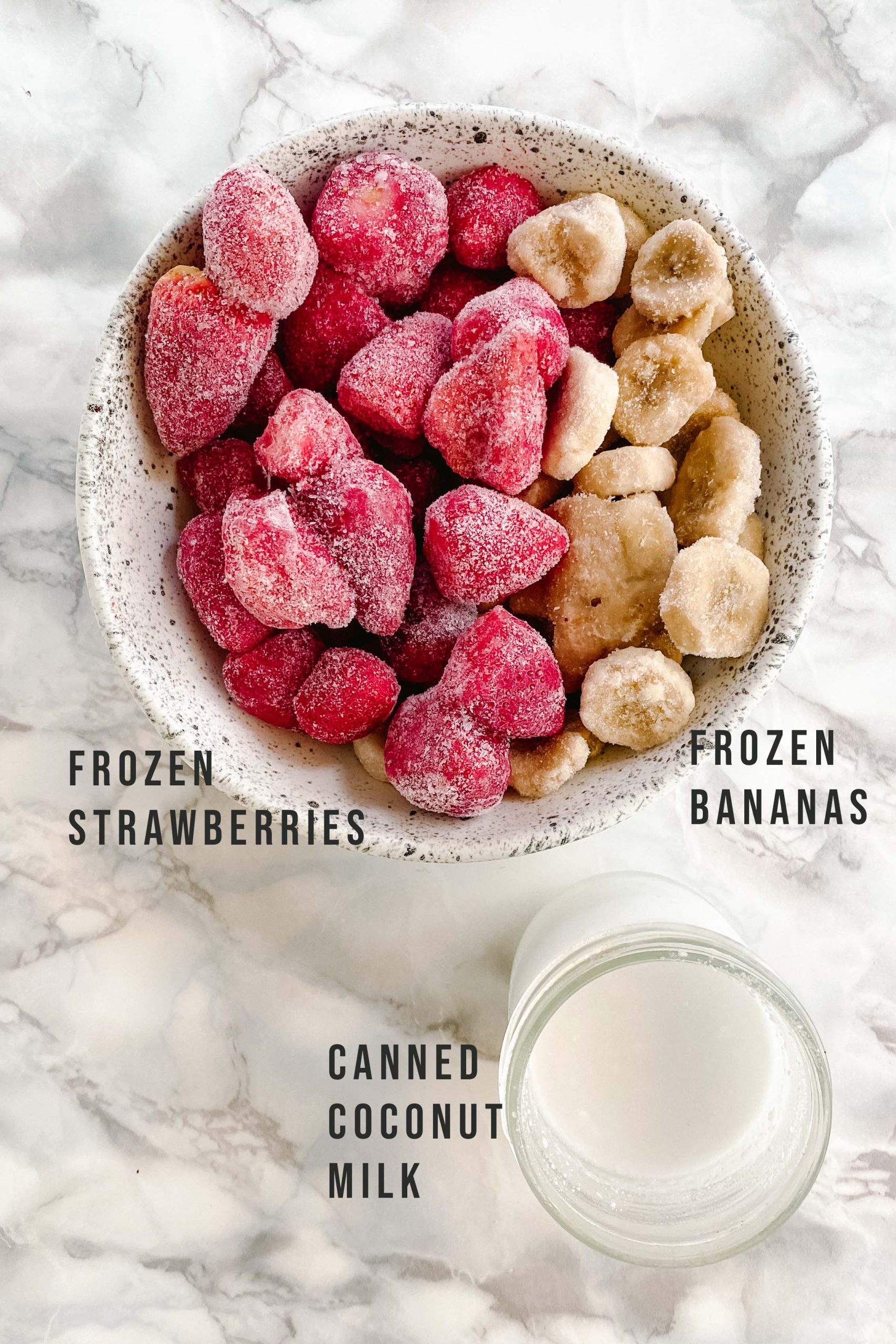A bowl of frozen strawberries and bananas beside a jar of coconut milk on a white marble counter.