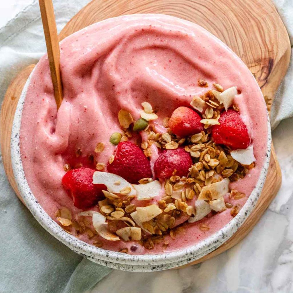 A pink smoothie bowl with strawberries and granola on top.
