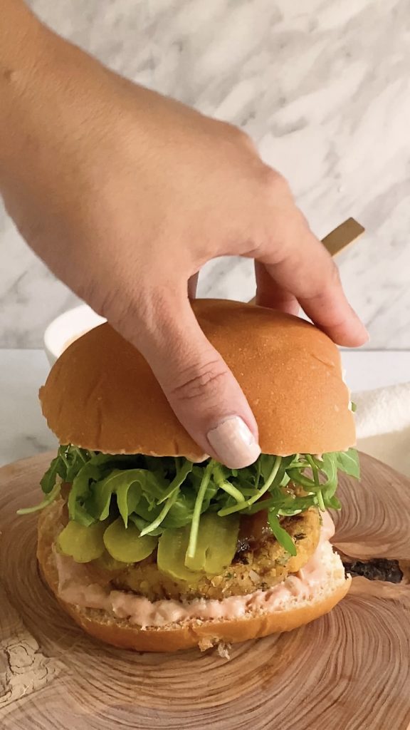 A hand adds a top bun to a topped burger.