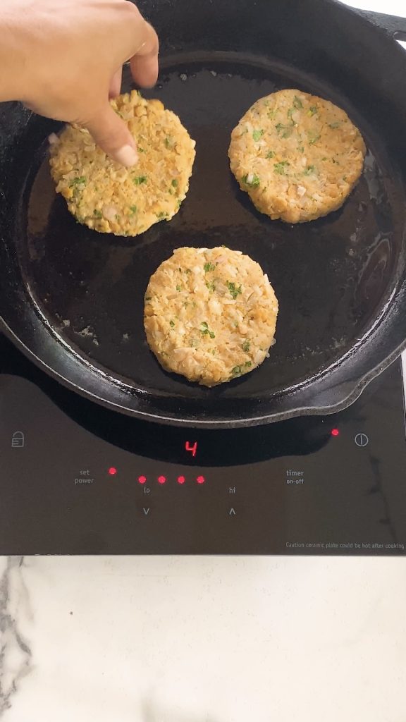 A hand places chickpea patties into a cast iron pan.