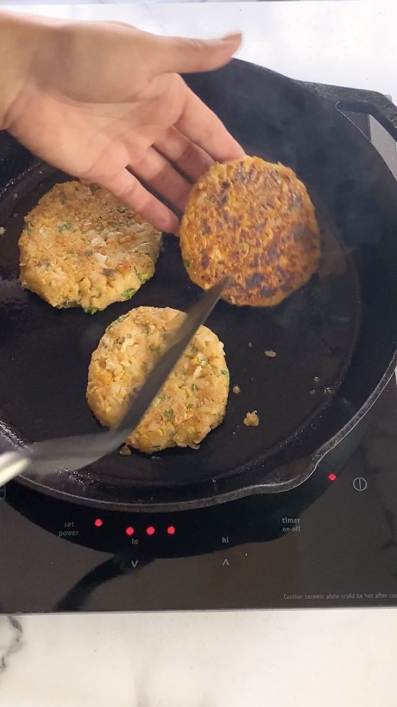 A hand helps a spatula flip a chickpea patty in a cast iron pan.