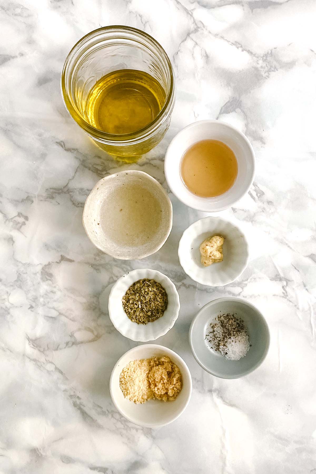 Ingredients for greek dressing in small bowls on a white marble counter.