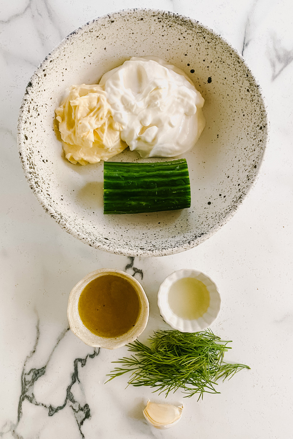 A bowl with mayo, yogurt, cucumber, a bowl with oil, a bowl with lemon juice, and dill and a clove of garlic on a white marble counter.