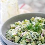 Chopped cucumbers in a bowl covered in tzatziki sauce, garnished with fresh dill with a text title for Pinterest.