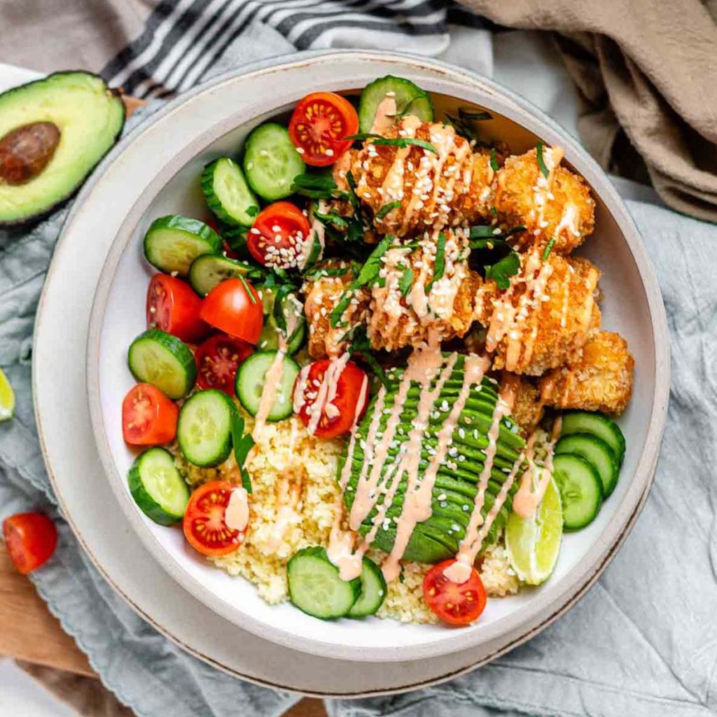 A bowl full of couscous, cucumber, tomato, avocado, and breaded tofu drizzled in sauce.