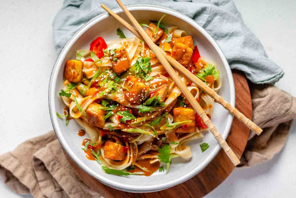 Spicy Peanut Tofu Bowl with rice noodles, red peppers, and creamy peanut sauce with chopsticks.