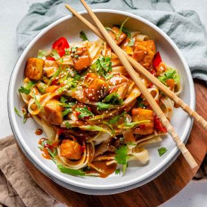 Spicy Peanut Tofu Bowl with rice noodles, red peppers, and creamy peanut sauce with chopsticks.