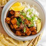 Tofu in sauce in a bowl beside rice, garnished with lemon slices and green onions.