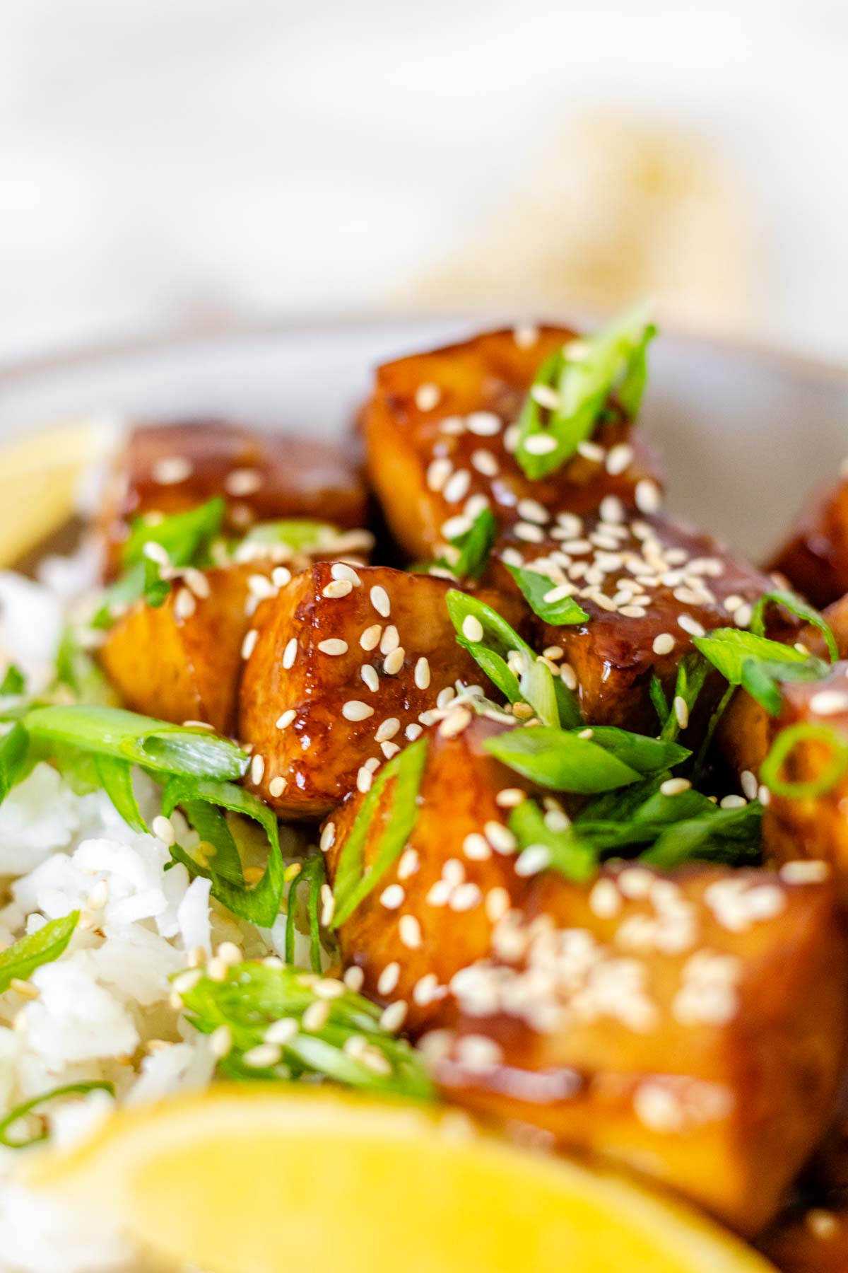 Tofu in sauce in a bowl beside rice, garnished with lemon slices and green onions.