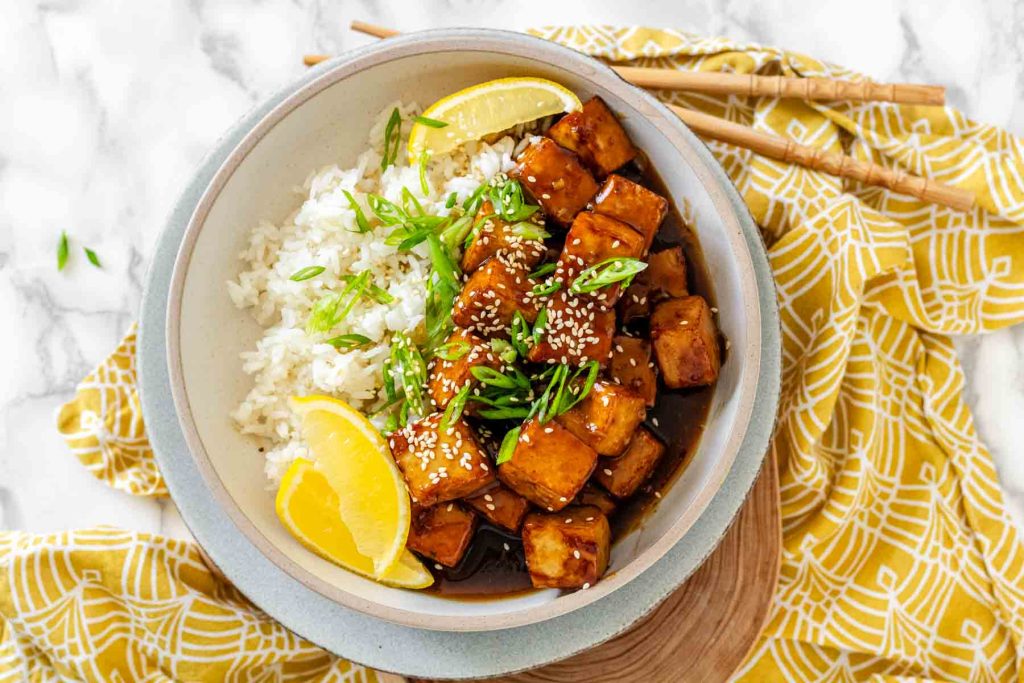 Tofu in sauce in a bowl beside rice, garnished with lemon slices and green onions.