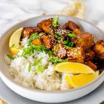 Tofu in sauce in a bowl beside rice, garnished with lemon slices and green onions.