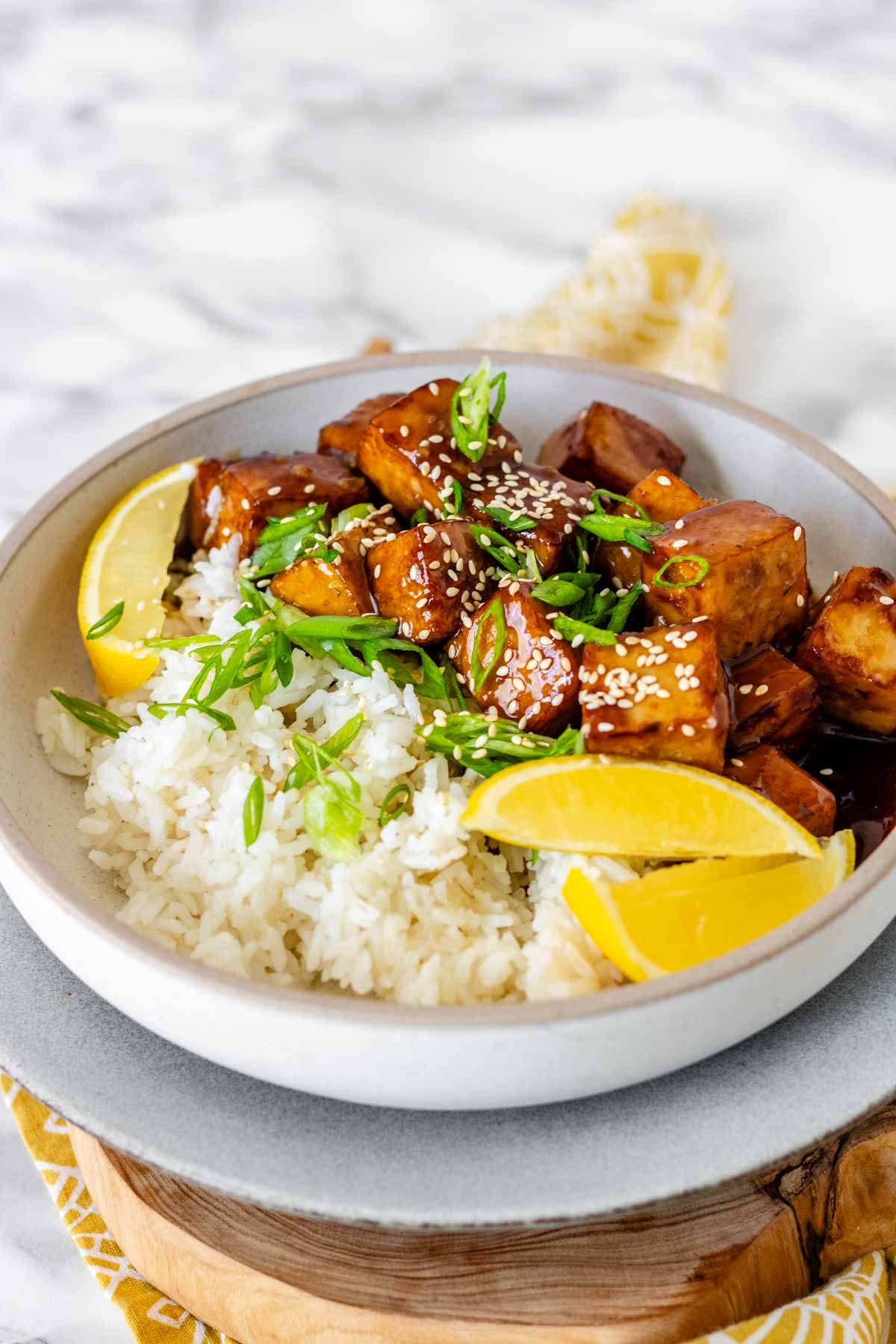 Tofu in sauce in a bowl beside rice, garnished with lemon slices and green onions.