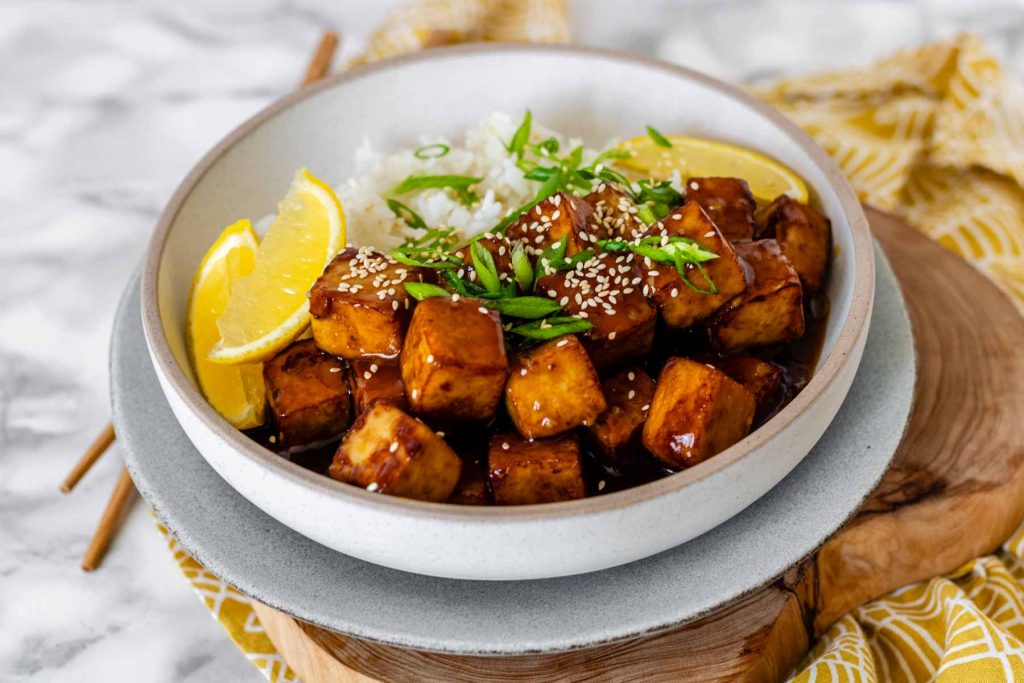 Tofu in sauce in a bowl beside rice, garnished with lemon slices and green onions.