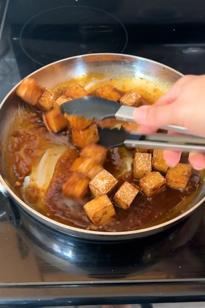 Tofu cubes tossed with tongs in a skillet of sauce.