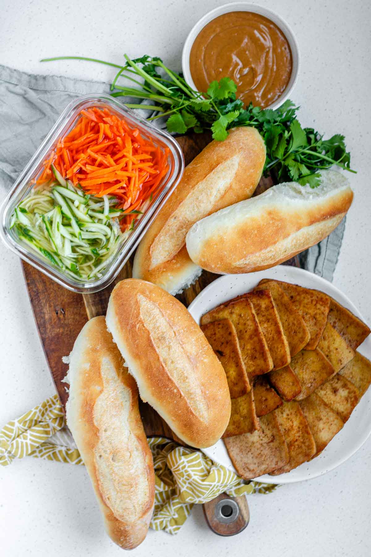 Ingredients for a sandwich, including sub buns, peanut sauce, cooked tofu, and pickled vegetables.